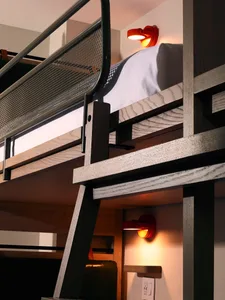A minimalist, industrial-style bedroom featuring a wooden bunk bed with cabinets underneath and a modern red lamp.