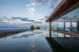 A modern rooftop swimming pool with a glass barrier overlooking a mountainous landscape under a cloudy sky.