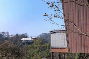 A modern, minimalist treehouse made of wood, elevated on stilts amidst lush greenery and forested mountains.