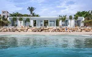 A modern beach house featuring glass walls and concrete columns, situated on a sandy shore with palm trees and blue ocean wat