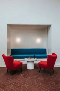 A modern living room with concrete walls and brick flooring, featuring a blue couch and red armchairs.