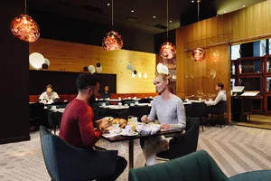 Modern restaurant interior with patrons seated at tables, contemporary lighting fixtures, and wooden walls.