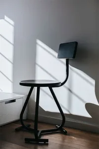 Modern black metal stool with a round seat and backrest, placed against a white wall with shadows from a window.