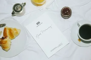 A minimalist breakfast setup featuring white linens, a croissant, black coffee, and a silver teapot on a white surface.