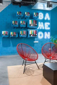 A modern lobby features red metal and fabric lounge chairs, a blue brick wall with neon sign reading "GAME ON", and a gray fl