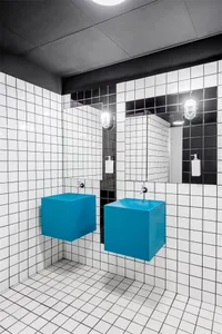A modern public bathroom features white and black grid-patterned tiles with bright blue square floating sinks and silver fauc