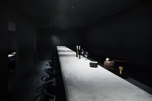 A minimalist restaurant bar with a concrete countertop and black seating on a dark tiled floor.