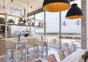 A modern restaurant bar featuring metal composite materials with a Happy Hour sign, high tables, and bar stools overlooking a