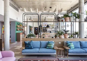 A modern office lobby featuring blue sofas with green and blue patterned cushions, concrete pillars, and potted plants.