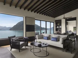 A modern living room featuring a white couch with blue cushions, a wooden floor, and ceiling beams, overlooking a scenic lake