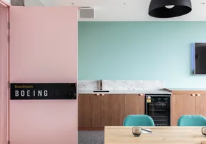 A modern office space featuring an industrial design with pink and green walls, wooden tables, and a kitchenette area.