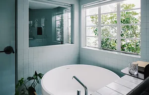 A minimalist bathroom with a white round bathtub, green plants, and blue-tiled walls near a window.