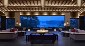 A modern hotel lounge featuring concrete and wood materials with a fireplace and seating areas by a large window overlooking 