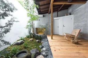 A serene Japanese-style patio with wooden deck, concrete walls, stone and gravel landscaping, and a wooden rocking chair.