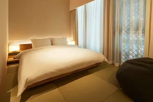 A minimalist bedroom featuring a white bed with wooden frame and fabric bedding, set against a neutral background with soft l