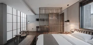 A minimalist bedroom featuring a wooden bed and furniture, with large windows and a hanging lamp.