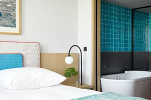 Minimalist bedroom with a bed featuring a wooden headboard and turquoise bedding, next to a bathtub adorned with turquoise ti