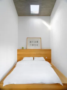 A minimalist bedroom with a wooden platform bed, white linens, and a concrete ceiling with a skylight.