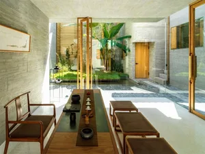 A modern living room featuring teak furniture, a small table with decorative items, and a view of a courtyard with greenery.