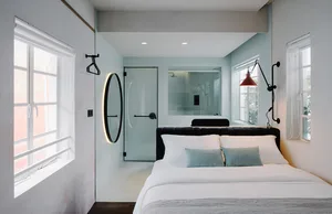 Modern minimalist bedroom featuring white concrete walls, glass shower enclosure, and a bed with white bedding and green acce