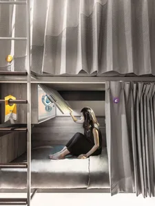 Person sitting on a minimalist bunk bed with a wooden frame and curtains.