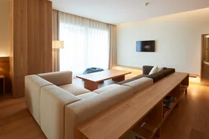 A modern living room with a wooden table, beige sofa, and a TV mounted on the wall, set against a light-colored wall and floo