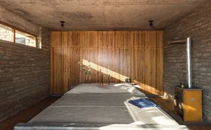 A minimalist bedroom with wooden furniture and a stone wall, featuring a concrete ceiling and a large window allowing sunligh