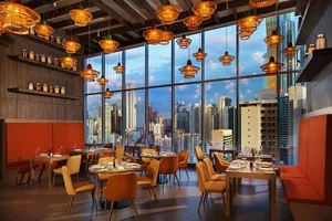Modern restaurant interior with orange and brown lighting, large windows overlooking urban skyline, and tables set for dining