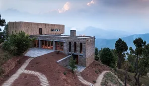 A modern house with stone and wood exterior, located on a hill with a view of the mountains and forest.