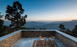 A terrace with stone walls and wooden chairs overlooking a mountainous landscape at dusk.