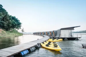 A floating house with a modern design, featuring glass walls and a wooden deck with kayaks.