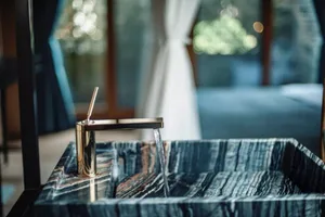 Water flowing from a golden tap into a marble sink, with a blurred background showing a white curtain and green plants.