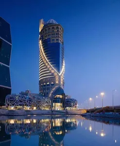 A modern, spiral-shaped building made of glass, concrete, and metal materials, reflecting in a nearby body of water.