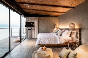 A minimalist bedroom with a white bed, wooden floor, concrete walls, and fabric accents.