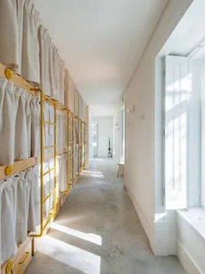 Minimalist hostel room featuring white curtains on bunk beds with yellow supports and concrete flooring.