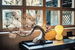 A modern wooden bonsai sculpture on a sleek black desk beside a yellow table lamp and a window with wooden frames.