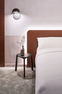 Minimalist bedroom featuring a brown upholstered headboard and white bedding on a bed with a black stool holding a small vase