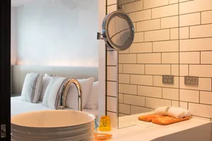 Modern bathroom with wood and white tiled walls, featuring a round magnifying mirror, a white sink, and a bed in the backgrou