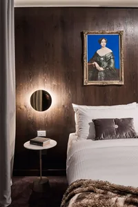 Modern bedroom featuring wooden panels, white bedding, a round table with a lamp, and a painting of a woman in a gold frame.