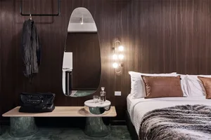 Modern bedroom featuring a wooden wall, a bed with white bedding, a round mirror, and a black leather bag on a side table.