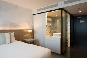 Modern minimalist hotel room with a white bed, glass door leading to a bathroom, and a wooden headboard.