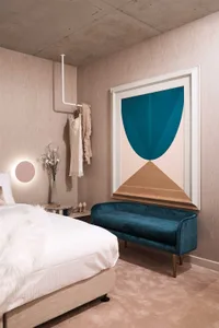 A modern minimalist bedroom with a blue velvet bench, geometric artwork, and a concrete ceiling.