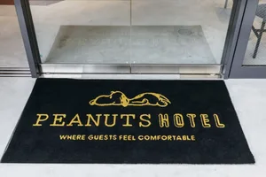 A black hotel door mat with yellow text that says 'PEANUTS HOTEL WHERE GUESTS FEEL COMFORTABLE'. The hotel logo features a sl