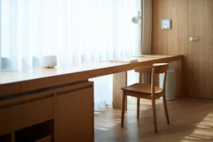 A minimalist home office space featuring wooden furniture, a window with white curtains, and a desk with a book.
