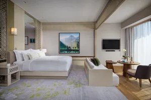 A minimalist hotel room featuring a bed with patterned cushions, a lounge chair, and a television mounted on the wall.