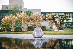 A woman meditates in the lotus position on a yoga mat in a serene urban garden park setting.
