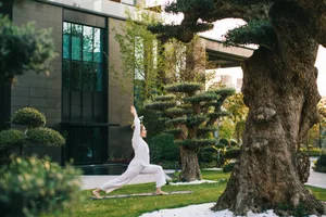 A woman performing a yoga pose in an urban garden wearing a white outfit, surrounded by green grass and trees.
