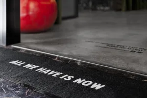 A black rug with the words 'ALL WE HAVE IS NOW' written on it, surrounded by a minimalist architectural space.