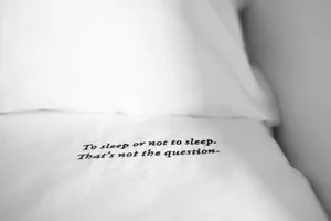 A white bed sheet embroidered with a famous Shakespearean quote, "To sleep or not to sleep. That's not the question."