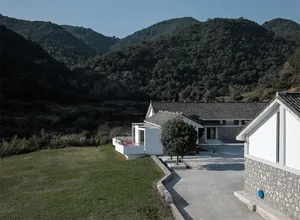 一棟擁有白色牆面與石材點綴的現代房屋，坐落於山坡上，周圍環繞茂密綠意。